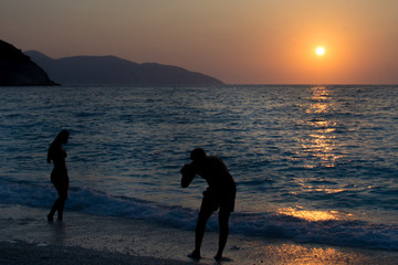 Silhouette man is taking pictures of a silhouette girl who is playing with waves at Myrtos beach in Kefalonia Greece. Sunset background