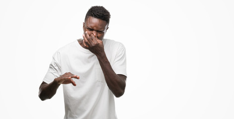 Wall Mural - Young african american man wearing white t-shirt smelling something stinky and disgusting, intolerable smell, holding breath with fingers on nose. Bad smells concept.