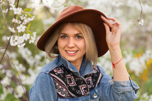 Blonde Hair Woman In Hippie Gypsy Jeans Jacket, Brown Hat Near White Tender Blossoming Tree. Fairy Dreamlike Mood Of Spring Or Summer. Beautiful Romantic Lady Smiling. Freedom Travel Lifestyle.