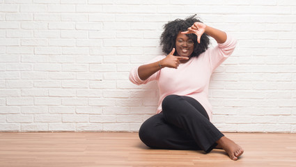 Poster - Young african american woman sitting on the floor at home smiling making frame with hands and fingers with happy face. Creativity and photography concept.