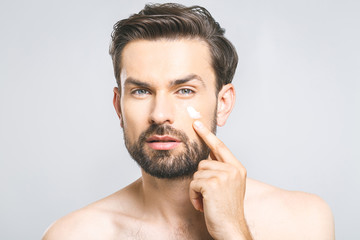 Skin care. Handsome young shirtless man applying cream at his face and looking at himself with smile while standing over gray background and looking at camera. Close-Up. Space for text.