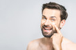 © denis_vermenko - Skin care. Handsome young shirtless man applying cream at his face and looking at camera with smile while standing over gray background and looking at camera. Close-Up. Space for text.