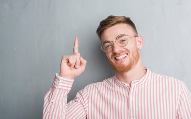 Wall Mural - Young redhead man over grey grunge wall surprised with an idea or question pointing finger with happy face, number one