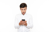 Fototapeta Na ścianę - Surprised man with short dark hair chatting or typing text message using cell phone, isolated over white background