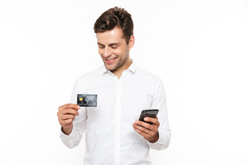 Wall Mural - Brunette happy man with dark hair holding smartphone and credit card, isolated over white background