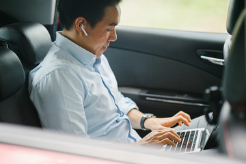 A young Japanese Asian man is talking on his smartphone using his bluetooth headset. He is riding at the back seat of a car he booked using a ride hailing app.