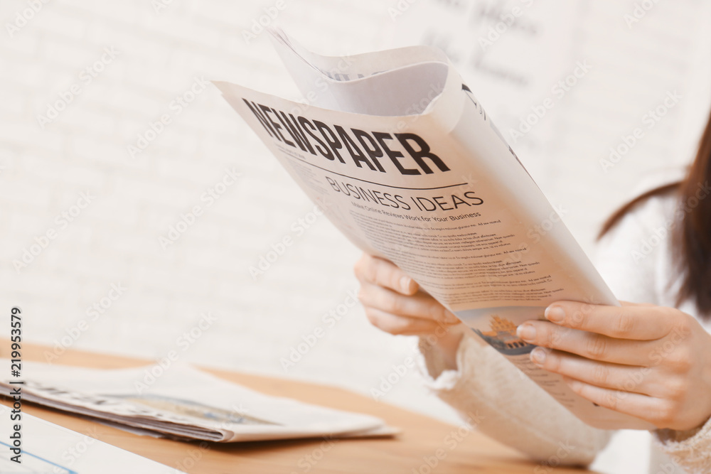 Young woman reading newspaper at table indoors