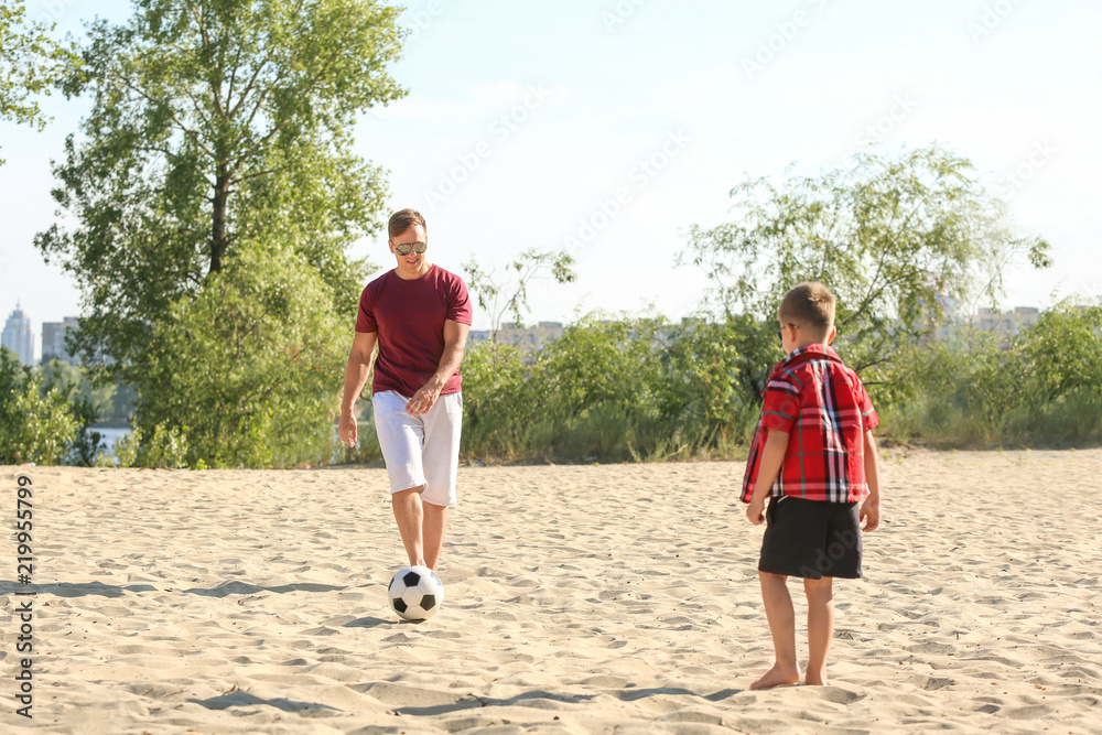 Little boy with his dad playing football on sand beach