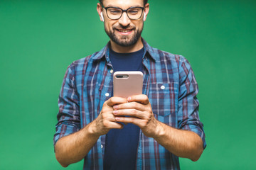 Wall Mural - Always in touch. Smiling young man holding smart phone and looking at it. Portrait of a happy man using mobile phone isolated over green background.