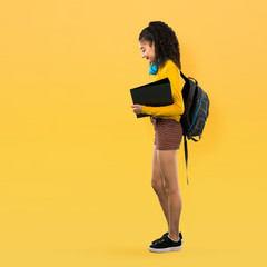 full body of teenager student girl with curly hair standing and looking down on yellow background