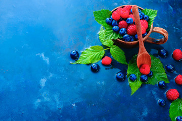 Colorful assorted mix of raspberry and blueberry with green leaves in a ceramic cup. Summer vitamins concept on a dark blue wet background with copy space