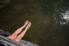 Young Woman Wearing Sandals Sitting On A Wooden Bridge, Hanging Her Legs Over The Water. Female Legs Hanging Over The River. Legs Hang From The Bridge Over The River With Seaweed.