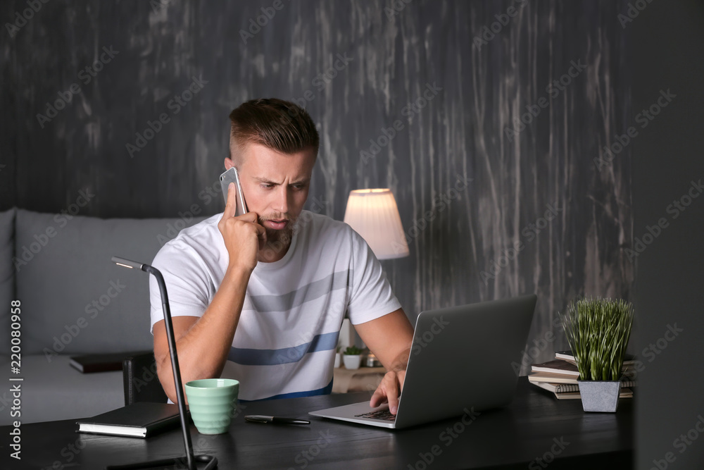 Young freelancer talking by phone while working on laptop in evening