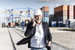 © Westend61 - Businessman at cargo harbor, wearing safety helmet, using smartphone