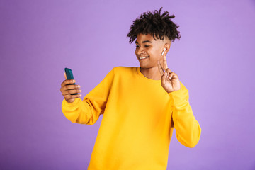 Poster - Portrait of young african american man smiling and taking selfie on smartphone, wearing bluetooth earpod, isolated over violet background