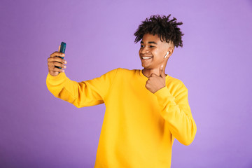 Poster - Portrait of teenage african american man smiling and taking selfie on smartphone, wearing bluetooth earpod, isolated over violet background