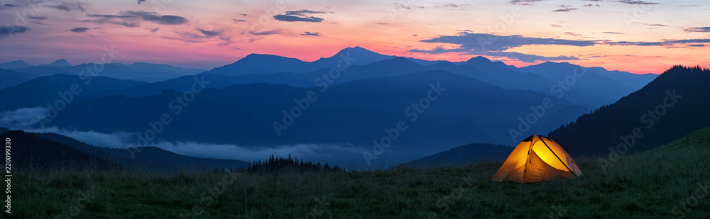 Glowing orange tent in the mountains under dramatic evening sky - obrazy, fototapety, plakaty 