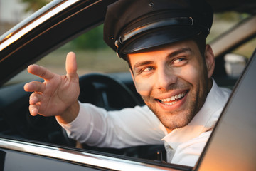 Portrait closeup of young caucasian chauffeur man wearing uniform and cap, smiling and greeting from car side window