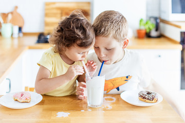 Children eating donuts and drinking milk on the white kitchen at home. Child is having fun with donuts. Tasty food for kids.