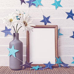 White chamomiles are in a gray vase. Nearby there is an empty wooden frame. On a white wooden background is a paper garland of blue stars.