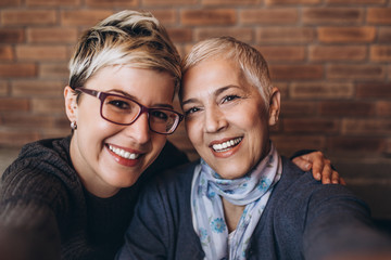 Wall Mural - Senior mother sitting in cafe bar or restaurant with her middle age daughter, they are smiling and taking selfie photo.