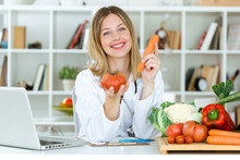Beautiful Young Nutritionist Looking At Camera And Holding Fresh Vegetables In The Consultation.