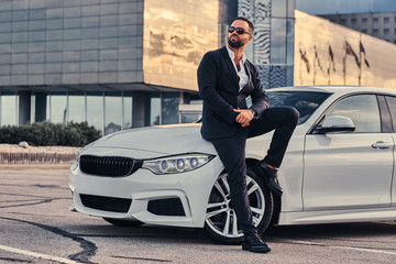 Handsome bearded male in sunglasses dressed in a black suit sitting on luxury car against a skyscraper.  