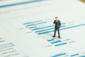 Investment, financial performance report analysis concept, miniature people figurine success businessman standing and thinking on printed stock exchange data graph and chart document