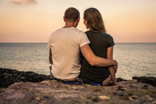 Man And Woman Hugging Sitting On Beach And Watching The Calm Sea At Sunset