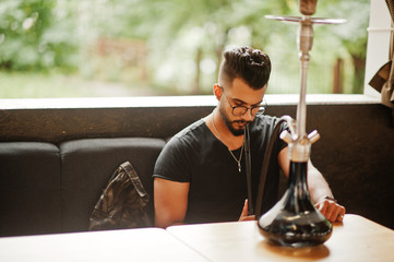 Stylish beard arabian man in glasses and black t-shirt smoking hookah at street bar. Arab model having rest.