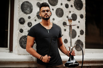 Stylish beard arabian man in glasses and black t-shirt smoking hookah outdoor. Arab model having rest.