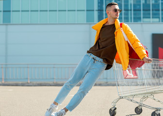 Wall Mural - fashion guy in sunglasses and a yellow jacket jump in a cart from food in the supermarket parking