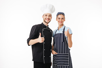 Wall Mural - Young smiling man chef in black uniform and white hat and pretty woman cook in striped apron and cap happily looking in camera while showing big thumbs up over white background