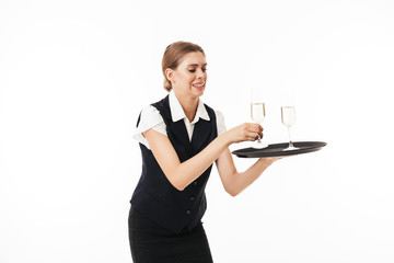 Wall Mural - Young waitress in uniform trying to hold tray with glasses over white background