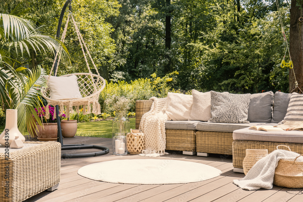 A big terrace with a comfortable leisure sofa with cushions, a table and a string swing in a green garden during sunny vacation. - obrazy, fototapety, plakaty 