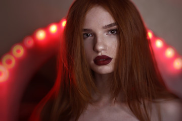 Concentrated young redhead lady with freckless and long hair looking at camera over red lighting background.