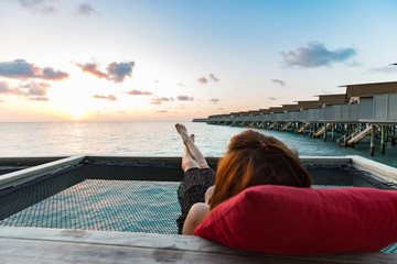 Back of view woman lying on terrace looking on sunset with villas over water in Maldives feeling relax and comfortable in Vacation summer holiday