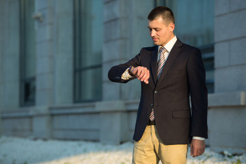 handsome young man in a suit standing near a building in the summer on the street looking at the clock