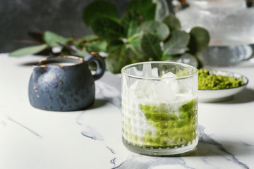 Matcha green tea iced latte or cocktail in glass with ice cubes, matcha powder and jug of milk on white marble table, decorated by green branches. Grey wall at background