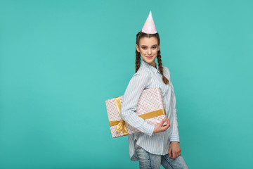 Wall Mural - Happy funny girl in striped blue shirt standing with dotted gift box and birthday cap on her head, looking at camera with safisfied smiley face, Indoor studio shot, isolated on green background