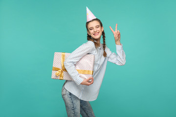 Wall Mural - Happy girl in striped blue shirt standing with dotted gift box and birthday cap on head, looking at camera with peace or victory sign and toothy smile, Indoor studio shot, isolated on green background