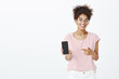 © Cookie Studio - Pick this phone model. Portrait of satisfied happy african-american woman in glasses and striped t-shirt, showing smartphone and pointing at device with index finger, smiling in approval expression