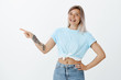 © Cookie Studio - Joyful carefree attractive woman in cropped top and jeans with braces and tattoos, holding hand on hip while pointing left with index finger, smiling broadly, being assured she showing right direction