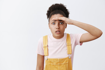 Wall Mural - Woman trying see through sick fog. Portrait of worried and confused attractive African American in yellow dungarees, holding palm near forehead and staring far away, standing over gray background