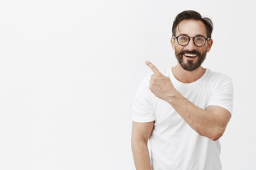 Happiness is just around corner. Portrait of charming friendly and carefree caucasian male with beard in glasses and t-shirt, pointing at upper left angle and smiling broadly over gray background