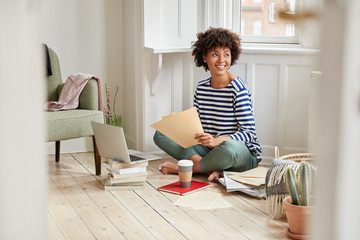 Satisfied student works on writing article for publication, dreams about holidays, sits crossed legs on floor against cozy domestic interior, surrounded with pile of books and modern laptop computer