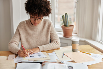 Young clever female student with Afro haircut makes graphic for economy lesson, works at home during weekend, dressed in warm oversized casual sweater, sits at desktop against window background