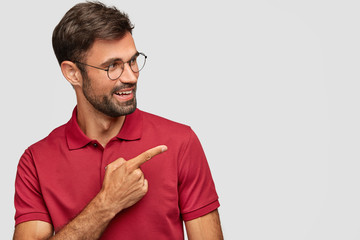 Wall Mural - Try that direction. Cheerful unshaven guy wears spectacles, shows way, points aside, has happy satisfied expression, wears red t shirt, isolated over white background, demonstrates free space