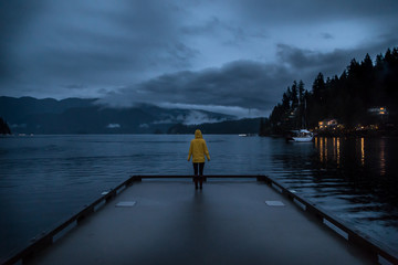 young woman standing in a yellow raincoat at the end of a pier with a view of the mountains and ocea