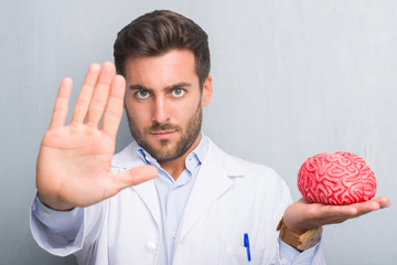 Wall Mural - Handsome young doctor psychologist man over grey grunge wall holding a brain with open hand doing stop sign with serious and confident expression, defense gesture